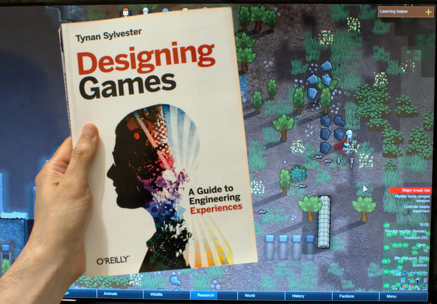 Book 'Designing Games', by Tynan Sylvester, being hold in front of a monitor displaying RimWorld.